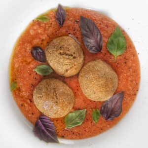 Tre finti canederli croccanti adagiati su una base di gazpacho di datterino e cetriolo, guarniti con foglie di basilico fresco.