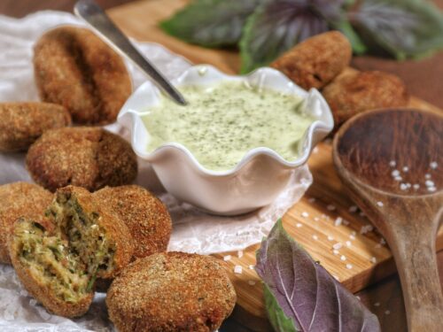 Polpettine di gamberi dorate servite con una salsa cremosa al basilico su un tagliere di legno.