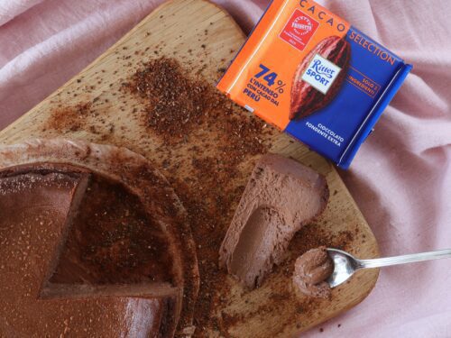 Una cheesecake al cioccolato tagliata a fette su un tagliere di legno, accanto a una tavoletta di cioccolato fondente Ritter Sport al 74% di cacao.