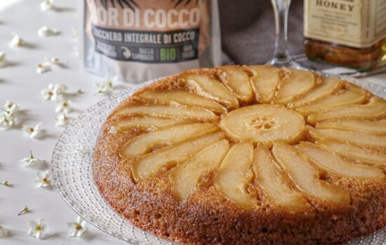 Una torta rovesciata alle pere disposta su un piatto di vetro decorato, con una confezione di zucchero di cocco e una bottiglia di liquore sullo sfondo.
