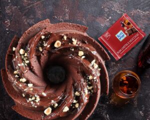 Una ciambella al cioccolato guarnita con glassa e granella di nocciole, accompagnata da una tavoletta di cioccolato Ritter Sport al rum e un bicchiere di liquore.
