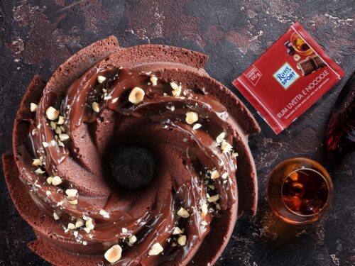 Una ciambella al cioccolato guarnita con glassa e granella di nocciole, accompagnata da una tavoletta di cioccolato Ritter Sport al rum e un bicchiere di liquore.