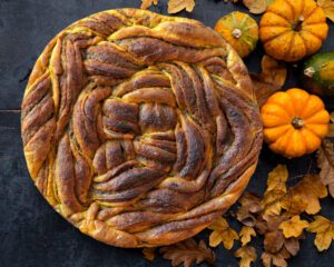 Una babka rotonda e intrecciata con una doratura scura, posata su un tavolo scuro accanto a piccole zucche decorative e foglie autunnali secche.