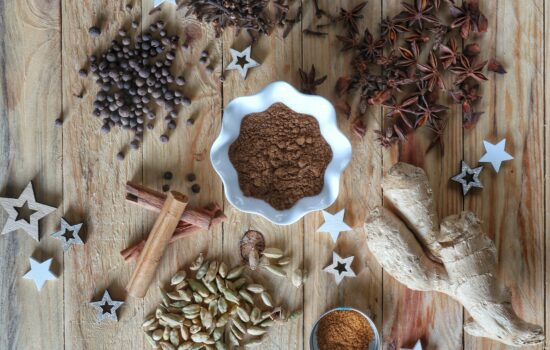 Una disposizione dall'alto di spezie aromatiche su un piano in legno, tra cui cannella, anice stellato, chiodi di garofano, cardamomo e zenzero, con una ciotola centrale contenente la miscela in polvere.