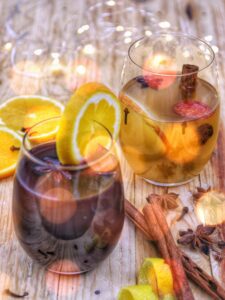 Due bicchieri di vin brulé, uno rosso e uno bianco, guarniti con fette di arancia e spezie su un tavolo di legno con luci soffuse.