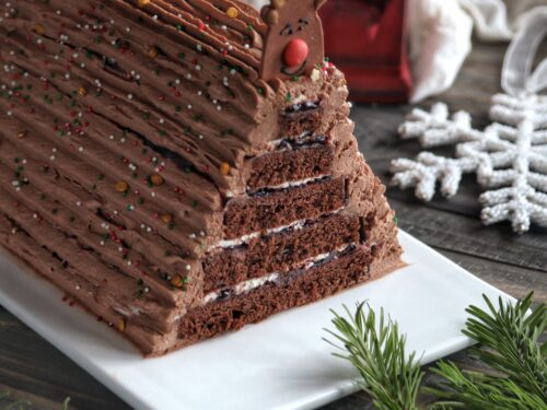 Torta a strati al cioccolato a forma di albero di Natale, decorata con crema al cioccolato, zuccherini colorati e una decorazione a forma di renna in cima.