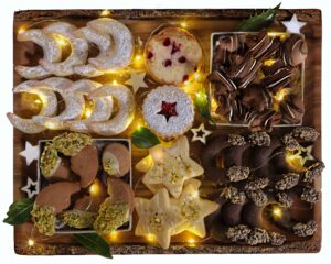 Un vassoio di legno decorato con luci natalizie contenente una varietà di biscotti artigianali, tra cui mezzelune allo zucchero, biscotti a forma di stella e dolcetti al cioccolato.