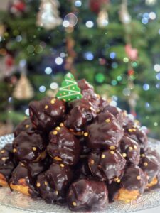 Un profitterol a forma di piramide ricoperto di glassa al cioccolato e decorato con un piccolo albero di Natale, su uno sfondo sfocato di luci natalizie.