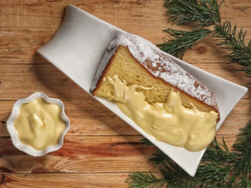 Una fetta di dolce soffice spolverata di zucchero a velo e ricoperta di zabaione tiepido su un piatto bianco, accompagnata da una ciotolina di zabaione.