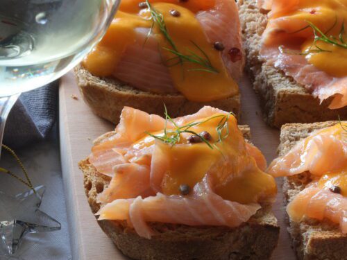 Crostoni di pane integrale guarniti con fette di salmone affumicato, salsa arancione, grani di pepe e aneto, serviti su un tagliere di legno accanto a un calice di vino.
