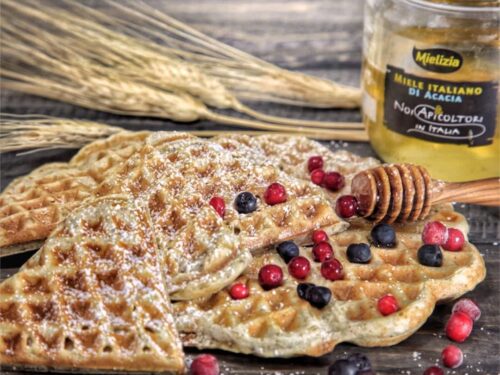 Waffel vegani guarniti con zucchero a velo e frutti di bosco freschi, accompagnati da un vasetto di miele di acacia Mielizia.