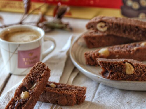 Tozzetti al cioccolato con nocciole disposti su un piatto e su un tovagliolo di lino accanto a una tazzina di caffè.