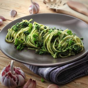 Un piatto di linguine condite con cime di rapa su un piatto scuro, poggiato su un tavolo di legno con spicchi d'aglio.