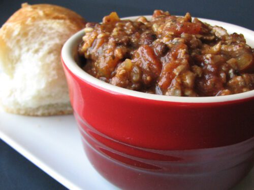 Una ciotola rossa contenente una pietanza densa a base di pomodoro e cereali, servita con un pezzo di pane su un vassoio bianco.