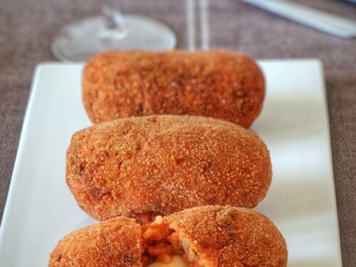 Tre supplì di riso dorati e fritti disposti su un piatto rettangolare bianco, con quello in primo piano aperto che mostra il cuore di mozzarella filante.