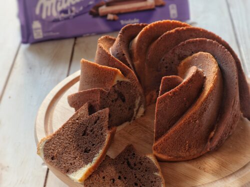 Una ciambella bicolore tagliata a fette su un tagliere di legno, con una tavoletta di cioccolato Milka sullo sfondo.