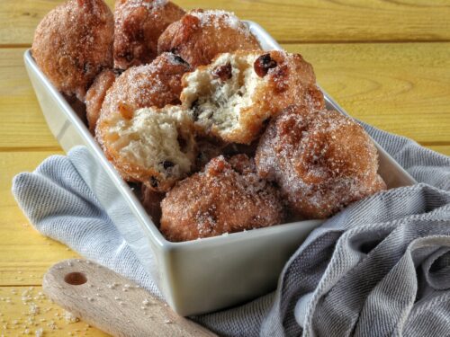 Un vassoio rettangolare bianco pieno di frittelle dorate all'uvetta ricoperte di zucchero semolato, appoggiato su un tavolo di legno giallo.
