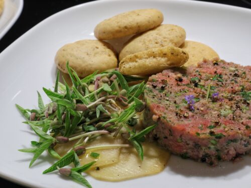 Un piatto di tartare di Scottona servito con fettine di cavolo rapa, germogli freschi e piccoli panini rotondi.