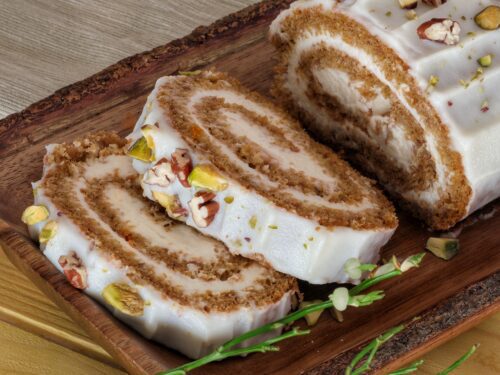 Un rotolo dolce farcito con crema bianca, glassato e decorato con granella di noci e pistacchi, servito su un vassoio di legno rustico.