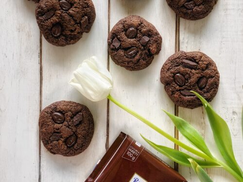 Biscotti al cioccolato con chicchi di caffè disposti su un tavolo di legno bianco accanto a un tulipano e una tavoletta di cioccolato Ritter Sport.