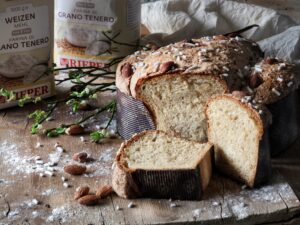 Una colomba pasquale tagliata su un tavolo di legno, circondata da mandorle, granella di zucchero e sacchetti di farina.