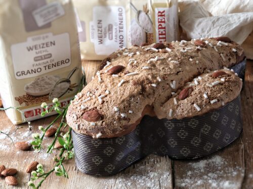Una colomba pasquale tradizionale decorata con mandorle e granella di zucchero, posizionata su un tavolo di legno accanto a sacchetti di farina.