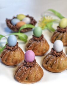 Piccoli dolci a forma di ciambellina guarniti con granella di cioccolato e un ovetto colorato, disposti su un piano bianco.