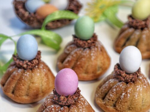 Piccoli dolci a forma di ciambellina guarniti con granella di cioccolato e un ovetto colorato, disposti su un piano bianco.