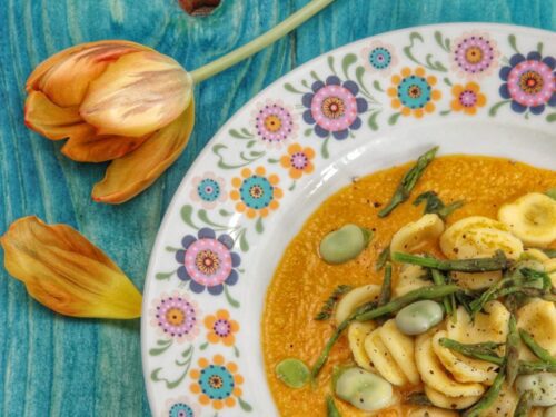 Un piatto di orecchiette condite con fave e bruscandoli su una crema arancione, servito in un piatto decorato con motivi floreali su un tavolo azzurro accanto a un tulipano.