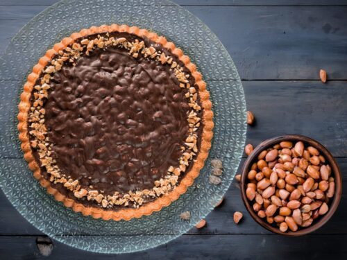 Una crostata al cioccolato guarnita con granella di arachidi su un piatto di vetro, accanto a una ciotola di arachidi intere.