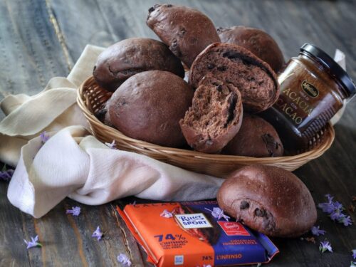 Un cestino di vimini pieno di pangoccioli al cioccolato scuro, accompagnato da un vasetto di miele al cacao e una tavoletta di cioccolato fondente Ritter Sport su un tavolo di legno.