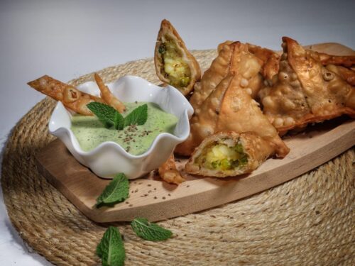 Samosa vegetariani dorati serviti su un tagliere di legno con una ciotola di salsa verde alla menta.