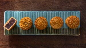 Cinque mooncake cinesi disposti in fila su un vassoio rettangolare di ceramica azzurra, con uno tagliato a metà per mostrare il ripieno scuro.