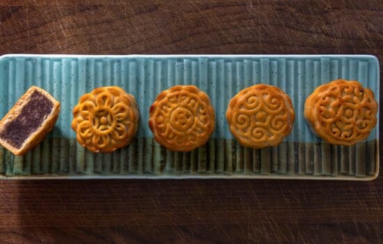 Cinque mooncake cinesi disposti in fila su un vassoio rettangolare di ceramica azzurra, con uno tagliato a metà per mostrare il ripieno scuro.