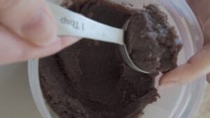Una mano utilizza un misurino da un cucchiaio per prelevare una porzione di pasta di fagioli rossi da un contenitore.