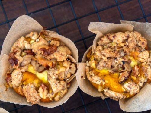 Due muffin dorati guarniti con noci e crumble, serviti in pirottini di carta su una griglia metallica.